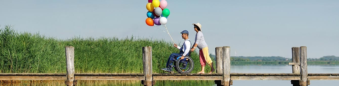 Mann im Rollstuhl hält eine große Traube mit Luftballons in die Luft, er wird von einer Frau über einen Steg geschoben