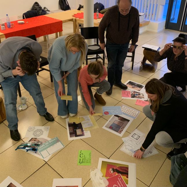 Mitglieder der Arbeitsgruppe bearbeiten Bilder und Bastelmaterial auf dem Boden.